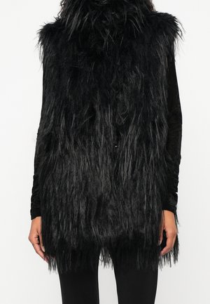 Personne portant une longue veste noire en fausse fourrure hirsute sur un haut noir à manches longues et un pantalon noir, debout devant un fond uni.