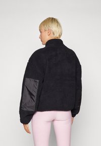 Sort fleece jakke med høj krave, der har kontrasterende glatte stofdetaljer på ærmerne. Tekstureret overflade med en afslappet pasform.
