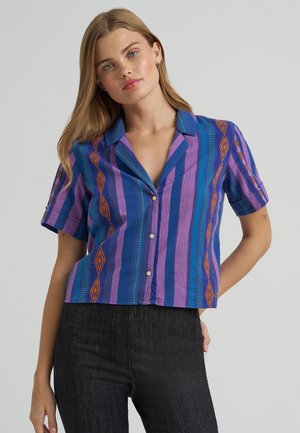 Mujer con cabello rubio que lleva una camisa de manga corta con botones y rayas verticales en púrpura, azul y naranja, junto con pantalones negros.