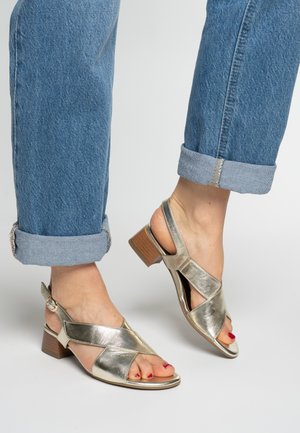 Metalen gouden sandalen met open tenen, gekruiste bandjes, lage houten hak en verstelbare enkellus, gedragen met opgerolde blauwe spijkerbroeken.