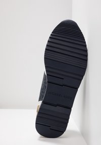 Semelle d'une basket noire avec des rainures profondes et le nom de la marque "Michael Kors" embossé sur la partie centrale texturée.