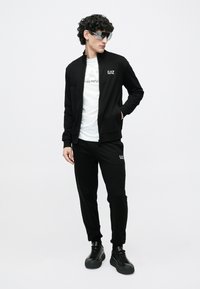 Μαύρο φόρμα που αποτελείται από ένα μπουφάν με φερμουάρ και αντίστοιχο παντελόνι, με λευκό λογότυπο στο μπουφάν και στο παντελόνι, συνδυασμένο με μαύρα αθλητικά παπούτσια.