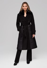 Blond kvinde iført en lang sort vinterfrakke med bælte og pelskrave, sorte bukser og sorte hæleboots, der står foran en grå baggrund.