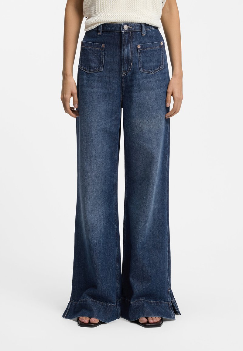 Jean en denim bleu foncé à jambes larges avec poches avant plaquées, taille haute et ourlets effilochés, porté avec des sandales noires à bouts ouverts et un haut en maille blanc.