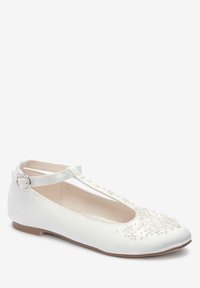 Chaussure plate blanche avec une sangle au niveau de la cheville et une boucle, ornée de petites appliques de fleurs 3D sur le bout et la sangle centrale.