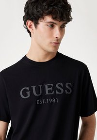 Musta puuvillainen t-paita, jossa on tekstuurilla varustettu harmaa broderie "GUESS" ja "EST. 1981." Pyöreä pääntie ja lyhyet hihat.
