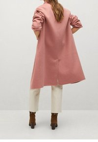 Lyserød uldblandingsfrakke med rullede ærmer, mellemlang design, slids på bagsiden, båret over cremefarvede bukser og brune hæle støvler, ensfarvet baggrund.