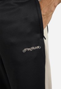 Sorte bukser med en cremefarvet sidepanel, prydet med det broderede "PEGADOR" logo i lys beige. Letvægtsstof, detalje med sidelommer.