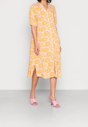 Vrouw met een gele bloemenjurk tot op de knie met knopen, pofmouwen, een zijsplit en roze hoge hakken slides.