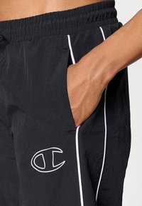 Crne sportske hlače s teksturiranim završetkom, bijelim akcentnim trakama uz bokove i izvezenim logotipom na prednjem džepu.