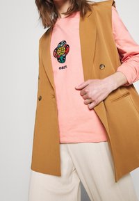 Tan bezrukávový blazer přes růžovou dlouhou košili s grafickým potiskem. Viditelné bílé plisované kalhoty. Knoflíková detailace na blazere.