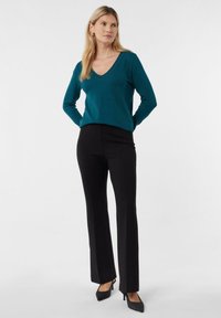 Kobieta ubrana w turkusowy sweter z dekoltem w serek, czarne proste spodnie i czarne szpilki, stojąca z rękami za plecami na jednolitym tle.