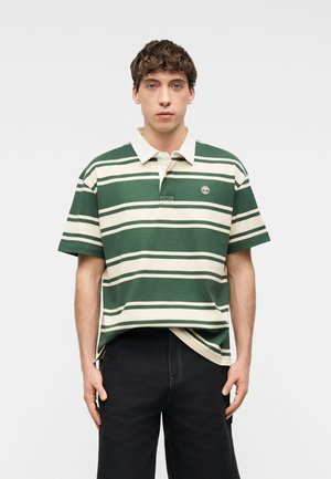 Fiatalember göndör hajjal, zöld és bézs csíkos galléros pólót és fekete nadrágot viselve, egyszerű világos háttér előtt áll.