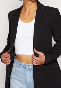 Černý přizpůsobený blejzr s uzavíráním na jedno tlačítko, nošený přes bílý krátký top a světle modré džíny. Hladká látka, strukturovaný design.