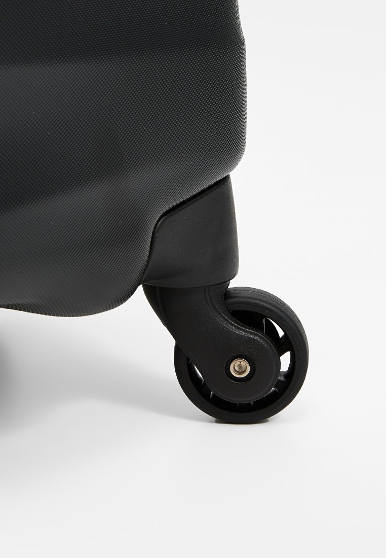 Valise à roulettes noire avec un corps texturé. Présente une roue noire ronde avec un moyeu argenté et un design robuste.