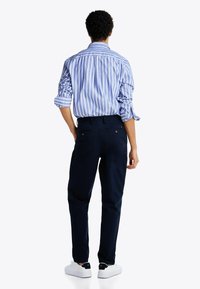 Camisa a rayas azul y blanca con mangas dobladas, combinada con pantalones de color azul marino oscuro. El modelo lleva zapatillas blancas y los pantalones tienen bolsillos traseros visibles.