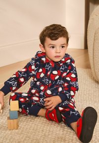 Czerwony i niebieski kapturek polarowy z nadrukiem Spider-Man, z białymi akcentami i kapturem. Pasujące czerwone kapcie Spider-Man. Siedzi na teksturowanym dywaniku.