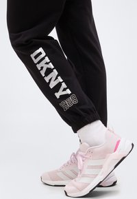 Άτομο που φοράει μαύρη φόρμα DKNY με το κείμενο «1989» και λευκές κάλτσες, συνδυασμένες με ροζ και λευκά αθλητικά Adidas σε λευκό φόντο.