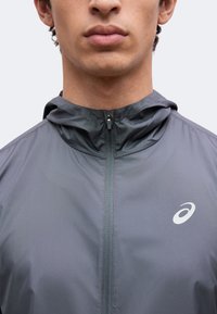 Jeune personne portant une veste grise zippée avec capuche et logo blanc sur la poitrine, montrée du cou au haut du torse sur un fond uni.