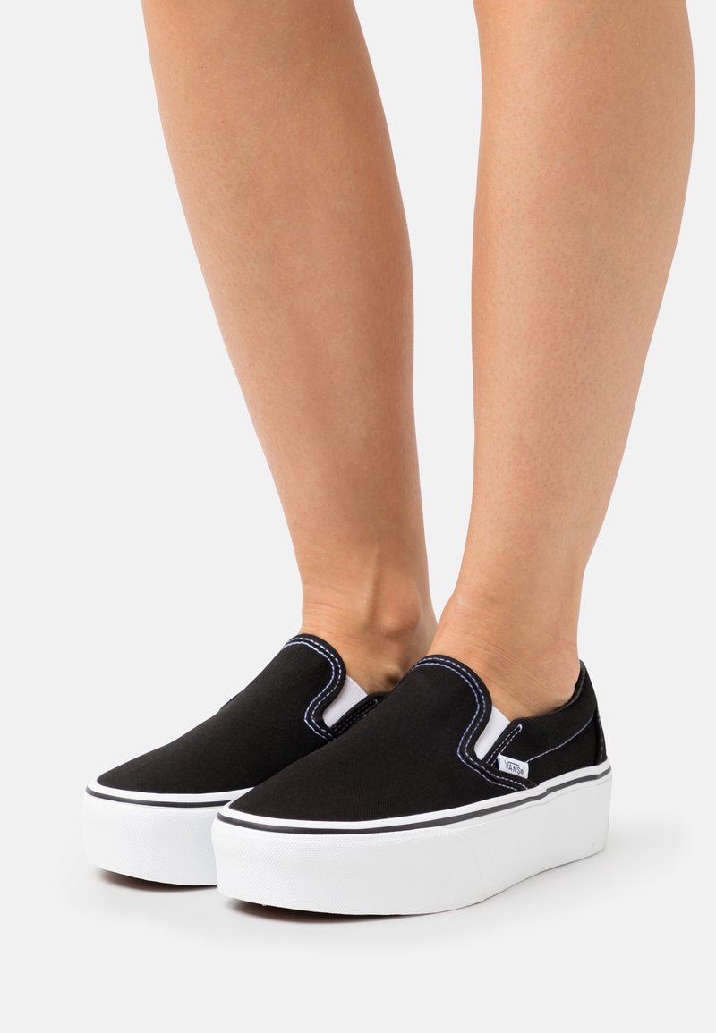Black canvas slip-on sneakers with a white rubber sole, navy stitching accents, and elastic side panels for easy wear.