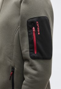 Κοντινό πλάνο μανικιού σακακιού με υφή ελιάς, με μαύρη τσέπη που διαθέτει κόκκινο φερμουάρ και το λογότυπο Icepeak γραμμένο με κόκκινα γράμματα.