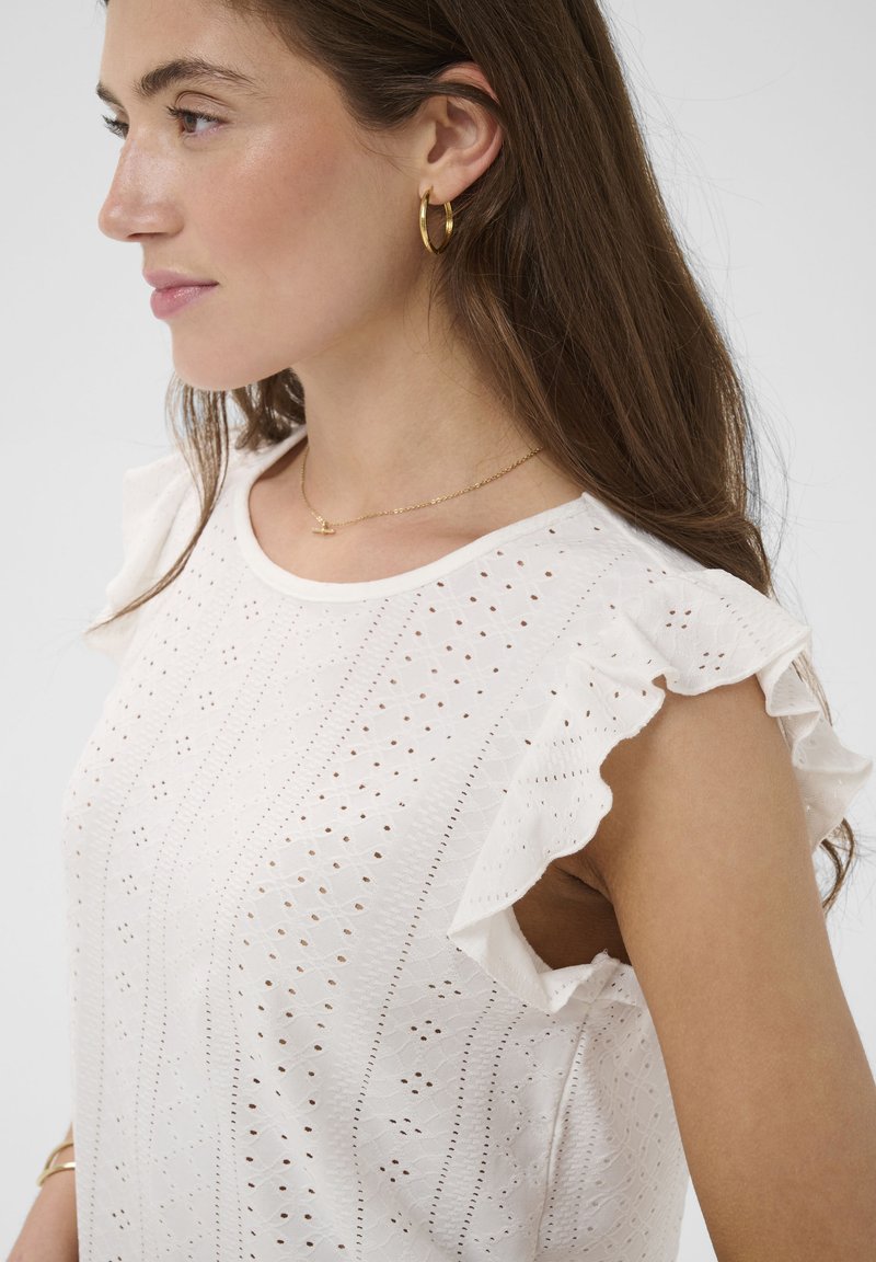Femme aux longs cheveux bruns portant des boucles d'oreilles créoles en or, un délicat collier en or, et un haut sans manches blanc à volants en broderie anglaise.