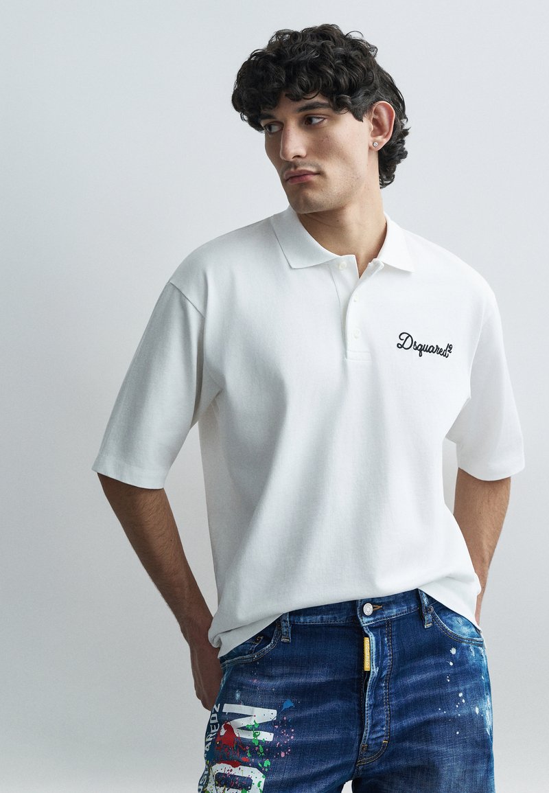 Polo blanca de manga corta con diseño de cuello, que presenta un logo bordado en negro en el pecho izquierdo. Usada con jeans azules estampados.