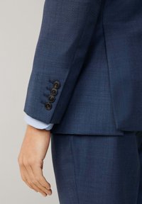 Marineblaues Sakko mit fünf dunklen Knöpfen an den Ärmeln, strukturiertem Gewebe und einem darunter sichtbaren hellblauen Manschette, die das strukturierte Design präsentiert.