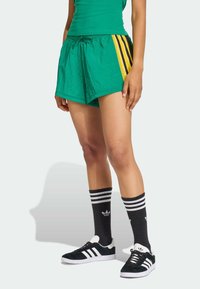 Gröna shorts med svarta och gula sidostriper, en texturerad yta, kombinerade med svartvita randiga strumpor och svarta sneakers.