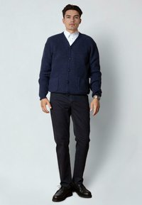 Man in een marineblauwe knooptrui, witte coll shirt, zwarte broek, zwarte schoenen en een horloge, staand tegen een effen lichte achtergrond.