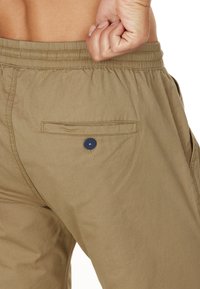 Beige Shorts mit einem elastischen Bund, ausgestattet mit einer Gesäßtasche, die durch einen marineblauen Knopf gesichert ist. Das Material wirkt weich und leicht.