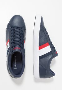 Navy leather sneakers with red and white striped accents. Features textured surface and round toe design. Lacoste logo on side. White sole.