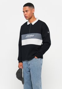 Jonge man in een zwart-grijze zip-up sweatshirt met lichte spijkerbroek, die een grijze baseballpet in één hand vasthoudt, staande tegen een witte achtergrond.