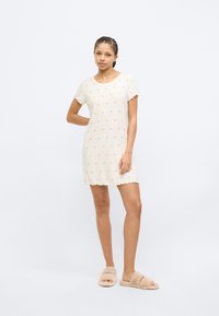 Σύντομο κρεμ νυχτικό με ροζ σχέδια καρδιών, με κυματιστό τελείωμα και μανίκια, κατασκευασμένο από ριμπ ύφασμα. Φορεμένο με απαλές μπεζ παντόφλες.