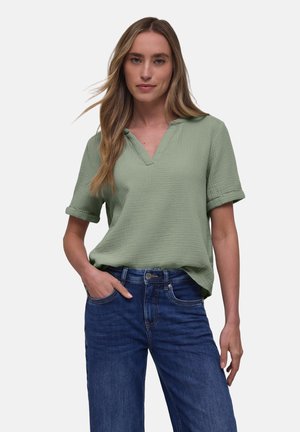 Kvinde iført en lysegrøn kortærmet bluse med V-hals og blå jeans, stående med den ene hånd i lommen.