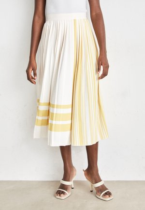 Woman wearing a pleated midi skirt with white and yellow vertical and horizontal stripes, paired with white high-heeled sandals.