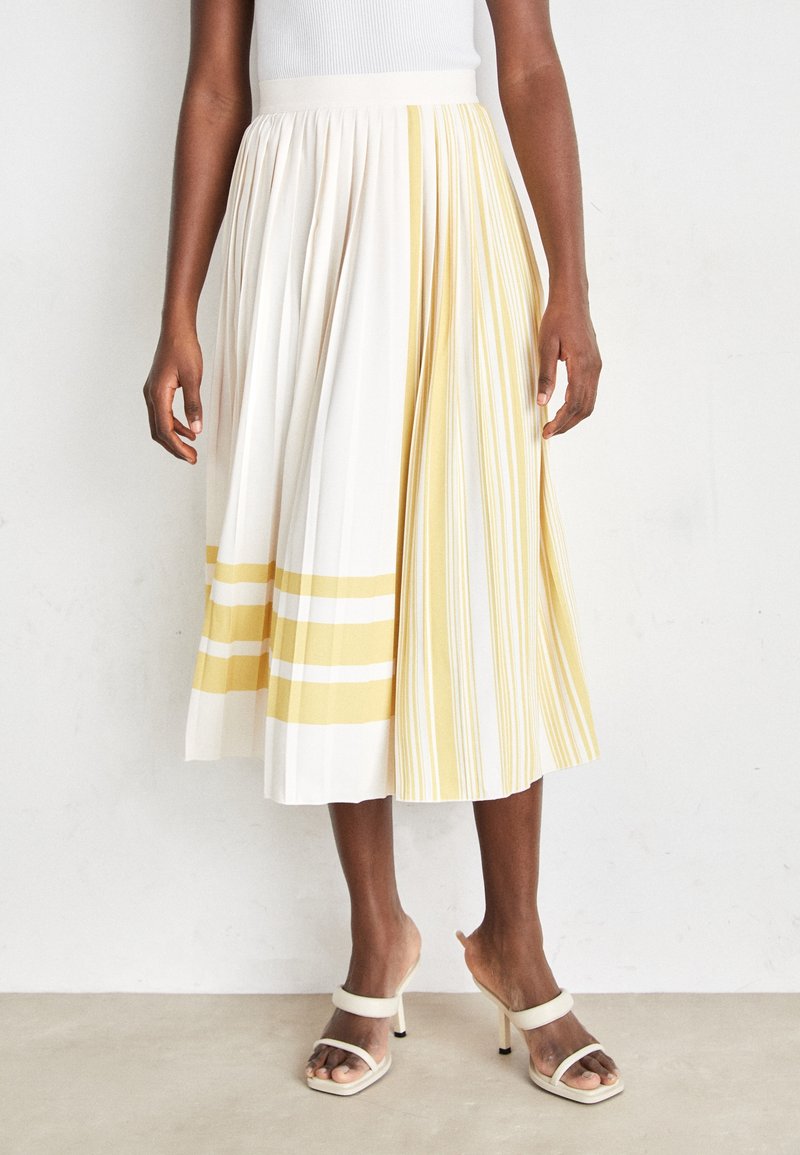 Femme portant une jupe midi plissée à rayures blanches et jaunes verticales et horizontales, associée à des sandales blanches à talons hauts.