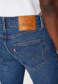 Jean en denim bleu avec une étiquette en cuir portant le design "Levi Strauss & Co." Coutures visibles et détails classiques des poches arrière.