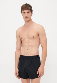 Young man standing against white background wearing black Calvin Klein boxer shorts, looking straight ahead with neutral expression.