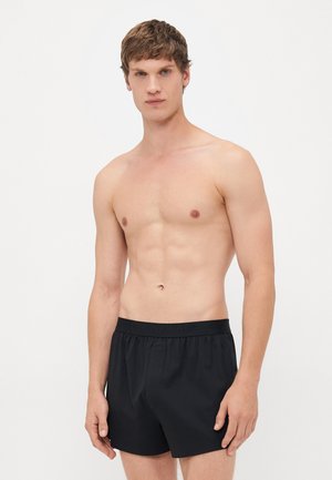 Junger Mann, der vor einem weißen Hintergrund steht und schwarze Calvin Klein Boxershorts trägt, mit neutralem Ausdruck geradeaus blickend.