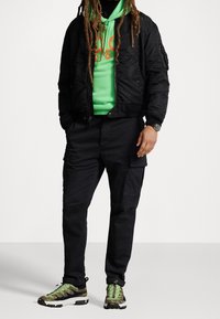 Svart bomberjacka över en ljusgrön hoodie med orange logotyp, kombinerat med svarta cargo-byxor och sneakers med gröna accenter.