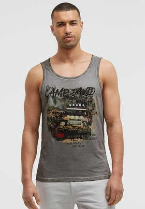 Mann trägt graues ärmelloses Shirt mit Geländewagen und Text über Abenteuer, steht vor einfarbig hellem Hintergrund.