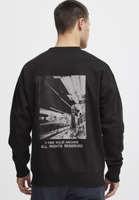 Sort sweatshirt med lange ærmer, prydet med et grafisk print på bagsiden af en metroplatform, samt teksten "© 1999 SOLID ARCHIVE."