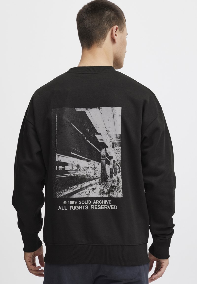 Sort sweatshirt med lange ærmer, prydet med et grafisk print på bagsiden af en metroplatform, samt teksten "© 1999 SOLID ARCHIVE."
