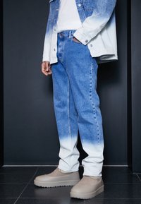 Osoba ve modrých ombre džínách, odpovídající denimové bundě, béžových slip-on botách, stojící na tmavém pozadí.