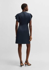 Marinblå klänning med kort ärm, figursydd midja och utsvängda ärmar. Slät tygkvalitet med en ren silhuett, i kombination med svarta remsandaler.