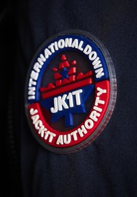 Cirkulär patch med blå, röda och vita färger. Har upphöjd text: "INTERNATIONAL DOWN JACK1T AUTHORITY" och grafiska element, inklusive en stjärna och snöflinga.