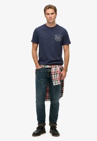 Camiseta de manga corta azul marino con un logotipo impreso, usada con vaqueros azules y una camisa de franela a cuadros atada alrededor de la cintura, zapatos negros.