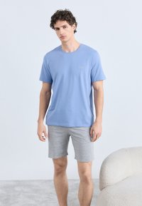 Camiseta de algodão azul clara com decote redondo e mangas curtas, combinada com calções cinzentos de algodão, numa superfície clara com carpete.