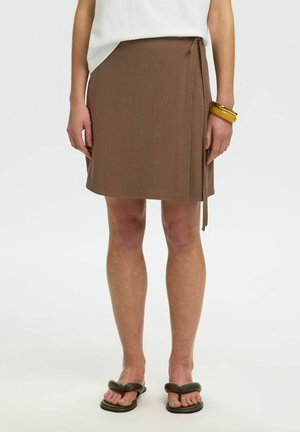 Femme portant une jupe portefeuille marron, un haut blanc, un bracelet jaune épais et des sandales noires rembourrées avec entre-doigts, posant devant un fond uni.